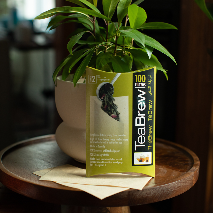 Translation missing: en.A box of TeaBrew paper filters stands on a small round wooden table next to a potted green plant. The box is olive green and black with white text, and a few beige filter papers are placed in front