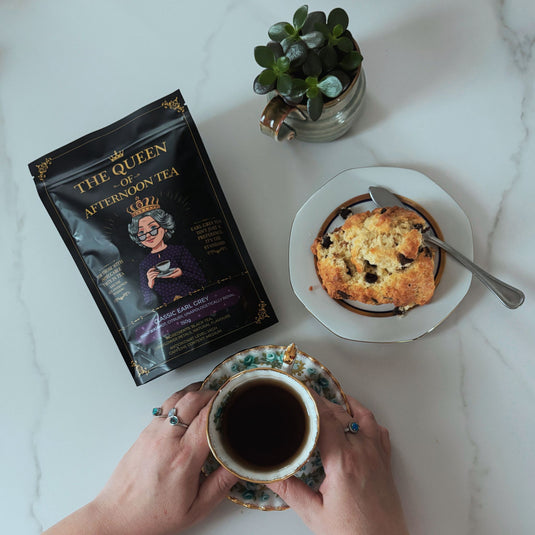 Translation missing: en.A top-down view of The Queen of Afternoon Tea holding a tea cup of black tea, with a bag of Classic Earl Grey tea next to it, and a scone on a plate on a white marble surface