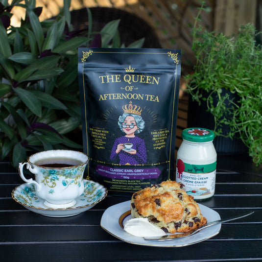 Translation missing: en.A package of Classic Earl Grey Tea from 'The Queen of Afternoon Tea' stands on a dark wooden table next to a cup of tea on a floral saucer, a scone with cream, and a jar of clotted cream. The scene is framed by green potted plants in the background