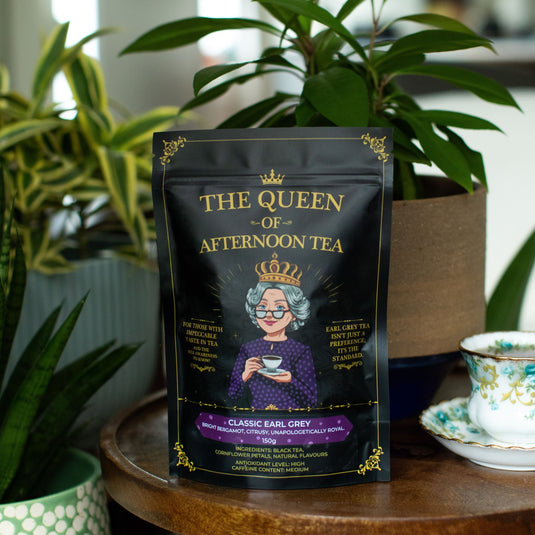 Translation missing: en.A stand-up pouch of 'The Queen of Afternoon Tea' Classic Earl Grey (150g) sits on a wooden table in a cozy, plant-filled setting with a teacup on the side