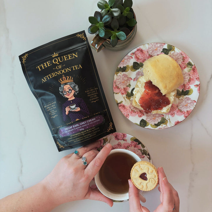 Translation missing: en.A top-down view of The Queen of Afternoon Tea holding a tea cup and a jammie dodger cookie, with a bag of Decaf Earl Grey Cream tea next to it, and a scone and jam on a plate on a white marble surface