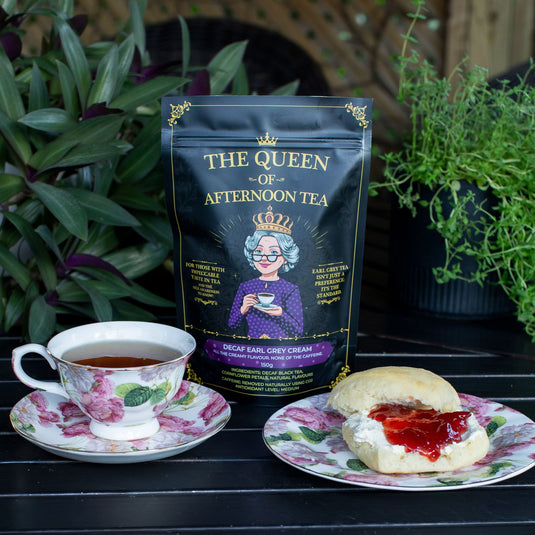 Translation missing: en.A stand-up pouch of Decaf Earl Grey Cream Tea by 'The Queen of Afternoon Tea' stands on a dark table between a floral teacup and saucer and a sliced biscuit with jam, with green plants in the background