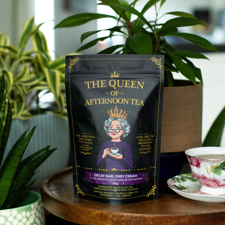 Translation missing: en.A stand-up pouch of Decaf Earl Grey Cream Tea by the 'The Queen of Afternoon Tea' stands on a wooden table in a styled tea-time setting, with a floral teacup and saucer to the right and potted plants in the background