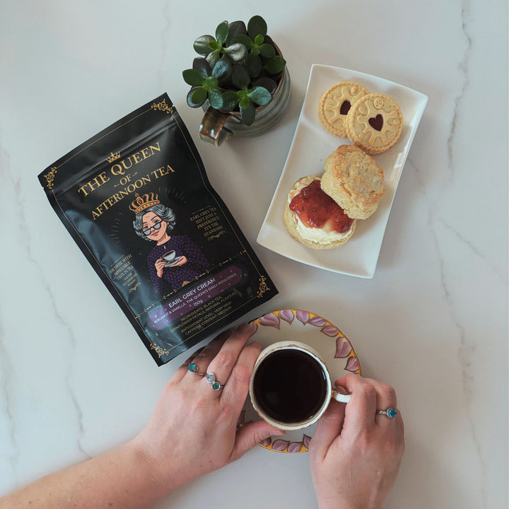 Translation missing: en.A top-down view of The Queen of Afternoon Tea holding a tea cup of black tea, with a bag of Earl Grey Cream tea next to it, and a scone and biscuits on a plate on a white marble surface