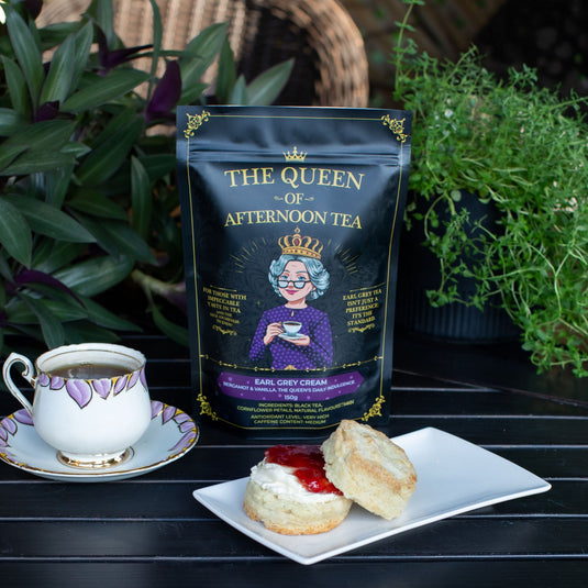 Translation missing: en.A stand-up pouch of Earl Grey Cream Tea from 'The Queen of Afternoon Tea' stands on a dark table beside a white teacup and a plate with a biscuit and cream. Green plants fill the background