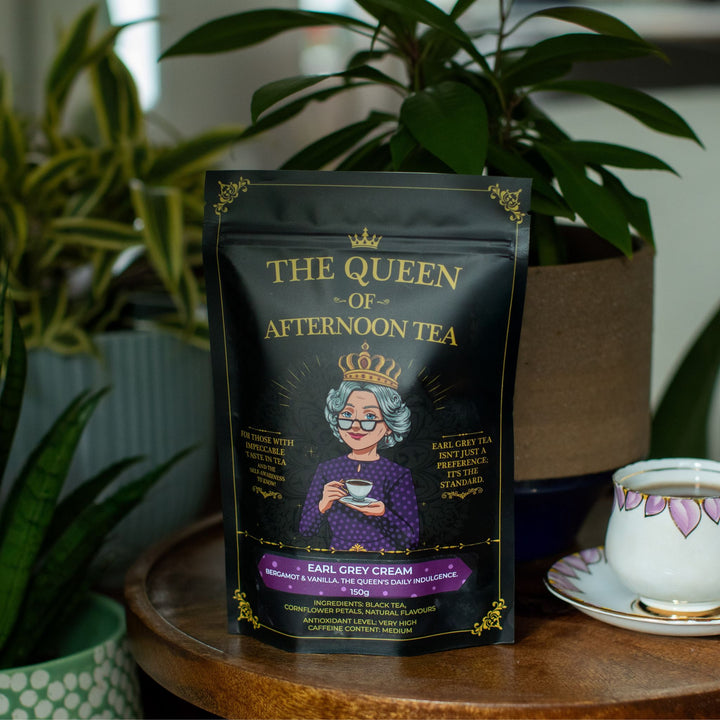 Translation missing: en.A stand-up pouch of 'The Queen of Afternoon Tea' Earl Grey Cream tea sits on a wooden table next to a teacup and saucer, with potted green plants in the background
