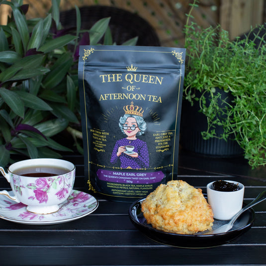 Translation missing: en.A black tea pouch labeled Maple Earl Grey by 'The Queen of Afternoon Tea' stands on a dark wooden table, styled with a floral teacup of tea, a scone, and a small dish of jam, with green plants in the background