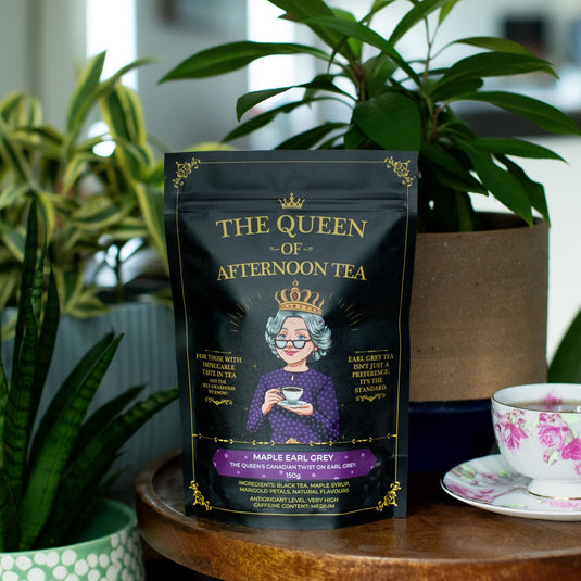 Translation missing: en.A stand-up pouch of The Queen of Afternoon Tea, Maple Earl Grey, is displayed on a wooden table in a styled home setting with potted green plants and a floral teacup on a saucer in the background